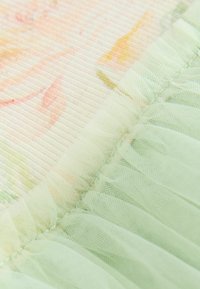 Close-up van een zachte doorschijnende groene stof met geplooide rand op een bleke bloemendruk textielachtergrond.