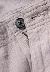 Lichtgrijs visgraatstof met een zwarte knoop, metalen ritsdetail en versterkte stiksels. Subtiele textuur en patroon zichtbaar.
