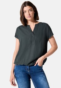 Mörkgrön kortärmad blus med V-ringning och avslappnad passform, tillverkad av lättviktsmaterial. Bärs med blå denimjeans.