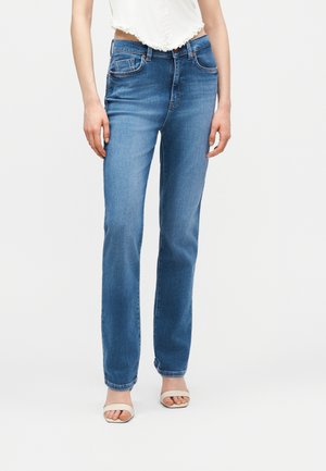 Γυναίκα που φοράει τζιν μεσαίας μέσης και ευθεία γραμμή σε μπλε χρώμα και λευκά σανδάλια ανοιχτά στημύτη, συνδυασμένα με λευκό κοντό τοπ.