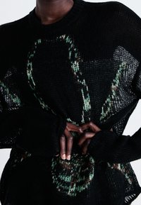 Zwarte gebreide trui met groen en bruin patroon. Heeft lange mouwen met duimopeningen en een losse, open geweven textuur.