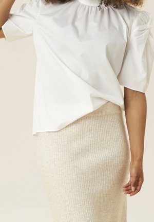 Femme portant un chemisier blanc à manches bouffantes rentré dans une jupe en tricot côtelée beige taille haute, un bras détendu le long du corps.