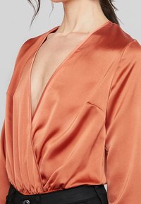 Satin-Oberteil in Rostfarbe mit tiefem V-Ausschnitt, langen Ärmeln und gerafftem Saum. Besitzt eine glatte Textur und fließende Drapierung.