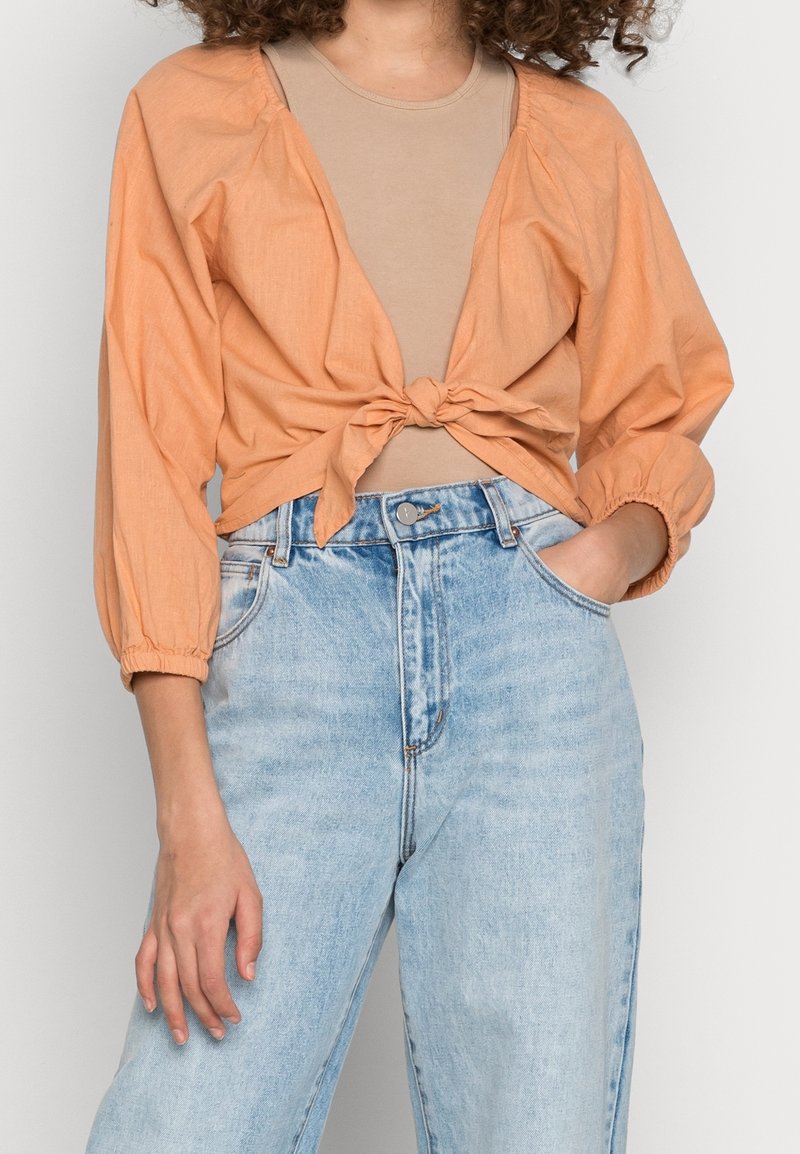 Blusa light orange con fiocco frontale e maniche a palloncino abbinata a jeans high-waisted blu chiaro. Tessuto morbido e vestibilità ampia.
