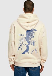 Felpa crema con un grafico blu sul retro che rappresenta una persona con le mani alzate, accompagnata dalla parola "Tupac" in un carattere stilizzato.
