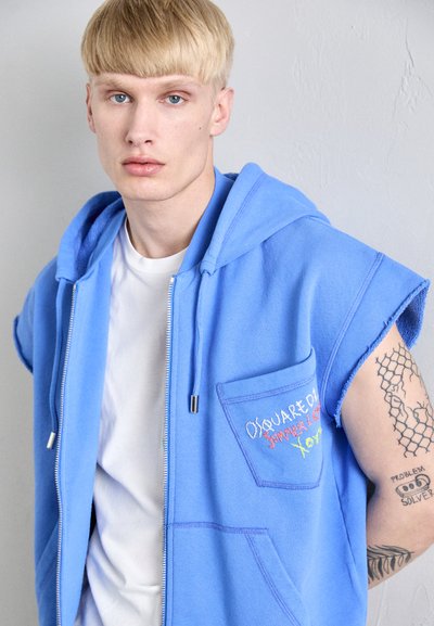 Blaues ärmelloses Zip-Hoodie mit Fronttasche und mehrfarbiger Stickerei; getragen über einem weißen Shirt, das einen strukturierten Stoff zeigt.