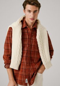 Camicia di flanella a quadri rossi e arancioni con bottoni marroni, indossata sotto un gilet in maglia a trecce color crema. Accenti di maglia testurizzata sulle spalle.