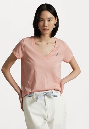 Pink cotton t-shirt with a v-neck, short sleeves, and a small blue logo on the chest. Worn with white pants and a light blue patterned belt.