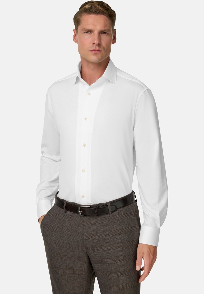 Uomo in camicia bianca con bottoni infilata nei pantaloni a strisce marroni, con una cintura nera, in piedi con una mano in tasca.
