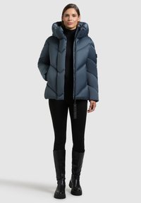 Pealisert sinine jope, millel on kõrge krae, diagonaalne tikandmuster, esikinnis tõmblukuga ja küljetaskud. Modell kannab musti pükse ja robustseid musti jalanõusid.