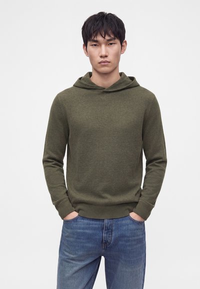Νέος άνδρας με κοντά μαύρα μαλλιά που φοράει μπλούζα με κουκούλα ελιάς και μπλε τζιν, στέκεται με τα χέρια στις τσέπες μπροστά από ένα απλό φόντο.