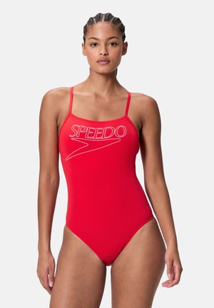 Junge Frau mit geflochtenem Haar trägt einen roten einteiligen Speedo-Badeanzug mit dünnen Schulterträgern vor einfachem Hintergrund.
