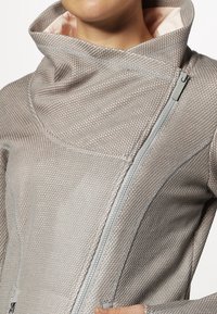 Giacca grigia chiara con texture, colletto alto e chiusura con zipper diagonale. Presenta cuciture evidenti e una fodera interna liscia.
