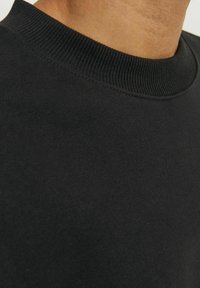 Detailní záběr na osobu v jednoduchém černém svetru s kulatým výstřihem, zobrazující límec a horní část hrudníku.