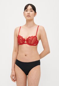 Raudonas permatomas nėriniuotas liemenėlė su dekoratyvine siūle ir lankeliais, turinti reguliuojamus dirželius, derinta su juodais pagrindiniais bikini kelnaitėmis.