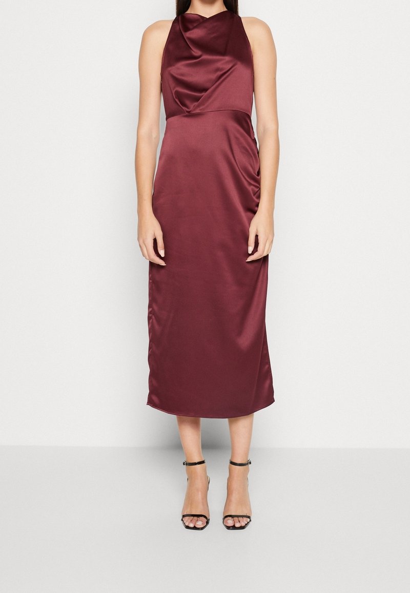 Femme portant une robe midi en satin bordeaux sans manches avec un col drapé et des sandales à talons hauts noires à bride à la cheville, debout contre un mur blanc.