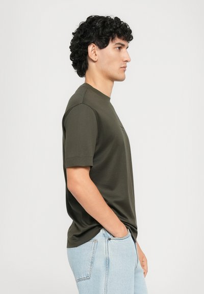 Fiatal férfi, göndör fekete hajjal, sötétzöld, rövid ujjú pólót és világoskék farmernadrágot visel, oldalról áll, egyik kezét a zsebébe dugva.