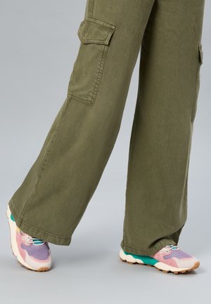Jambes portant un pantalon cargo ample vert olive et des baskets aux couleurs pastel avec des accents roses, violets et turquoise sur un fond gris.