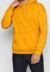 Gul hættetrøje lavet af blødt stof, med en forlomme foran, snøre i hætten og ribbede manchetter. Modellen har mørke jeans på.