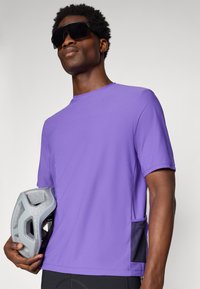 Camiseta deportiva corta de manga en color púrpura con cuello redondo y bolsillo lateral, acompañada de un casco gris que el modelo sostiene en la mano.