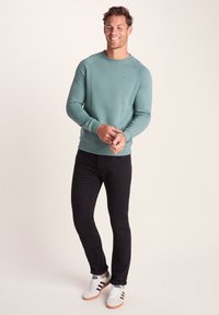 Green sweatshirt with a round neckline and ribbed cuffs, paired with black trousers and white sneakers featuring black stripes.