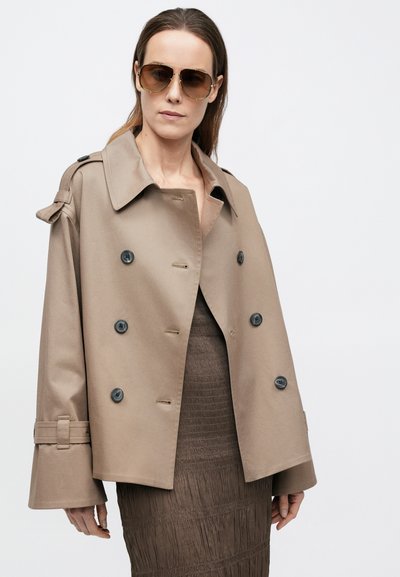 Beige trenchcoat met een dubbele rij knopen en zwarte knopen. Heeft brede mouwen en schouderepauletten, gedragen over een getextureerde bruine jurk.