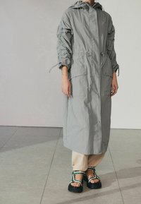 Lichtgrijze lange jas met getailleerde taille, capuchon met trekkoord en zakdetails; gestyled met beige broek en robuuste zwarte sandalen.