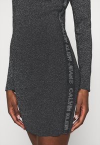Robe noire à côtes en tissu texturé, dotée de manches longues et d'un détail logo sur le côté en gris portant la mention "CALVIN KLEIN JEANS."