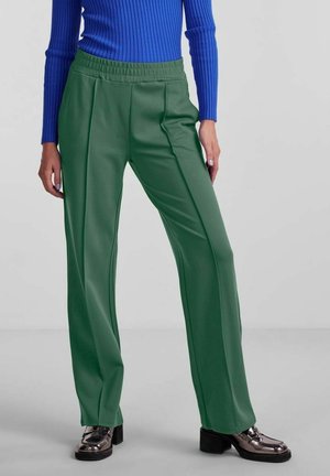 Groene, ruime broek met een gladde textuur en elastische tailleband, gecombineerd met een blauwe geribde top en glanzende zwarte schoenen.