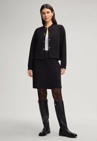 Veste noire courte avec des boutons dorés, top blanc, jupe noire, collants à motifs et bottes en cuir noires jusqu'aux genoux. Finitions mates et texturées.