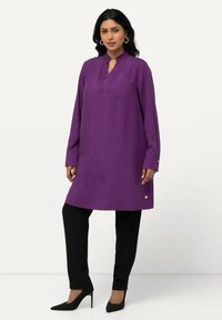 Paarse tuniek met lange mouwen, staande kraag en smockdetails aan de voorkant, gecombineerd met zwarte broeken en puntige zwarte hakken.