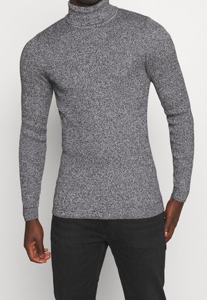 Homme portant un pull à col roulé gris chiné ajusté et un pantalon noir, debout les mains détendues le long du corps.