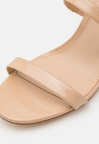 Sandale en cuir verni beige avec une bride frontale unique et une bride à la cheville, présentant des coutures détaillées et un bout carré.