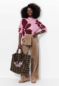 Un pull rose avec de grandes fleurs marron, un pantalon en velours côtelé beige, et un sac fourre-tout à carreaux avec du texte. Des bottes beiges chunky complètent la tenue.