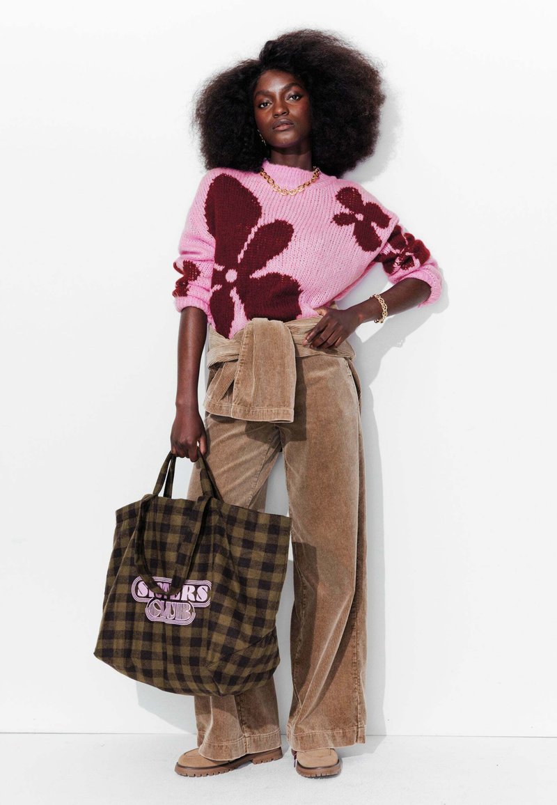 Un pull rose avec de grandes fleurs marron, un pantalon en velours côtelé beige, et un sac fourre-tout à carreaux avec du texte. Des bottes beiges chunky complètent la tenue.