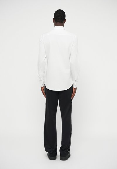 White long-sleeve shirt with a smooth texture, paired with black pants featuring side stripes and a relaxed fit. Back view only.