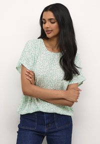Top verde chiaro a maniche corte con un motivo astratto bianco, caratterizzato da un collo rotondo e una vestibilità morbida, abbinato a jeans blu scuro.