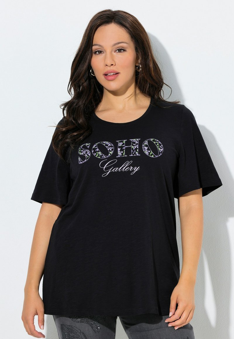 Donna con capelli lunghi e scuri che indossa una maglietta nera a maniche corte con la scritta floreale "SOHO Gallery", in piedi contro una parete bianca.