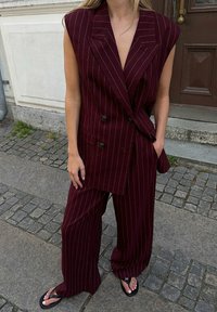 Burgundski brezrokavni blazer s črtami in dvojni gumbi, v kombinaciji s širokimi hlačami. Tkanina izgleda teksturirana, v paru z črnimi japonkami.