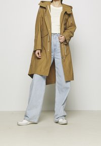 Beżowy wodoodporny płaszcz z kapturem, regulowanymi ściągaczami i przednimi kieszeniami, w połączeniu z jasnoniebieskimi, luźnymi jeansami i białymi trampkami.