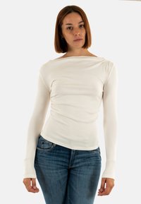 Maglietta bianca a maniche lunghe con scollatura a barca, caratterizzata da un design aderente con una leggera texture arricciata nella parte frontale, indossata con jeans blu.