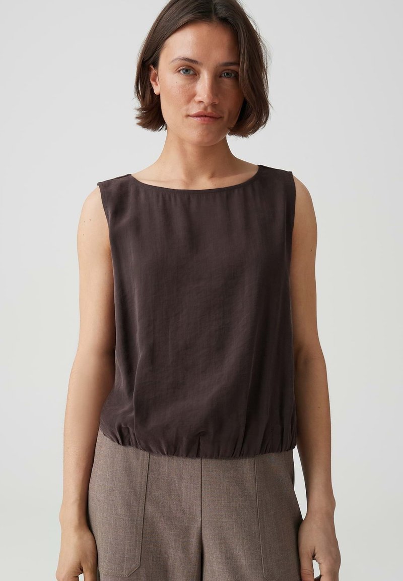 Woman with short brown hair wearing a sleeveless dark brown top and high-waisted light brown pants standing against a plain light background.
