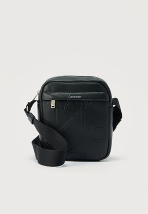 Bolso bandolera negro de Calvin Klein con patrón de logo en relieve, cremallera plateada y correa de tela ajustable sobre un fondo neutro.