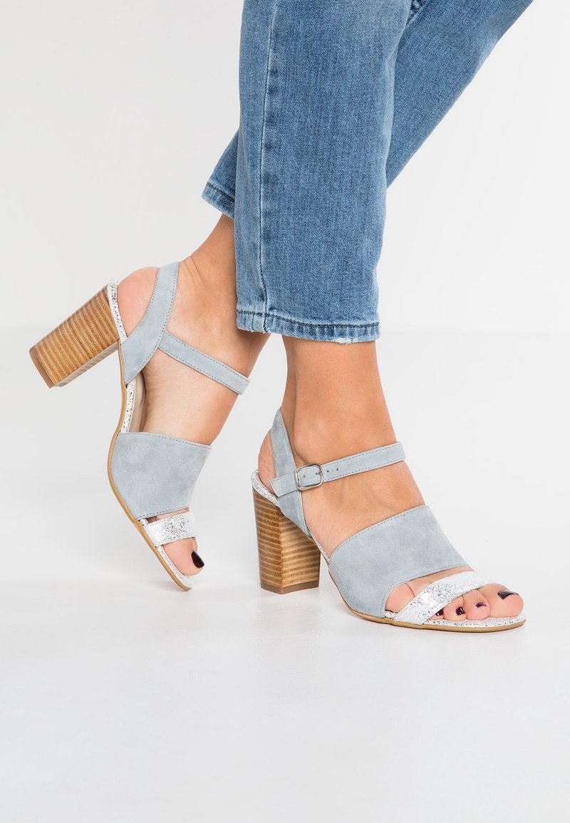 Sandales à talons hauts en daim gris avec un talon bloc en bois. Elles présentent une lanière argentée sur les orteils et une bride réglable à la cheville.