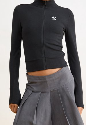 Frau trägt ein eng anliegendes schwarzes Adidas Zip-up Langarmshirt und einen hoch taillierten grauen plissierten Rock, Mittelteil sichtbar.