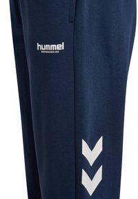 Granatowe spodnie sportowe z białym logo, bocznymi kieszeniami i charakterystycznymi białymi akcentami w kształcie chevronu na dolnej części nogawek. Miękki materiał bawełniany.