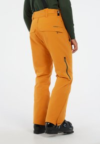 Oranje skibroek met verstelbare banden, zijzakken met rits en versterkte zoom, gecombineerd met zwarte skischoenen, tegen een witte achtergrond.