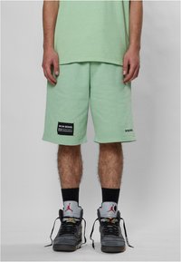 Shorts in cotone verde menta con una vestibilità rilassata, decorati con toppe nere del marchio e tasche laterali. Indossati con calze nere e scarpe da ginnastica grigie.