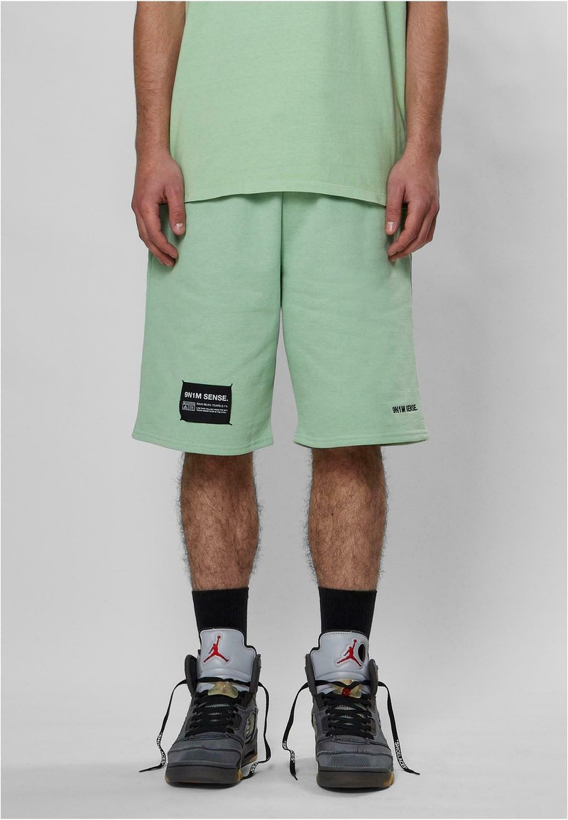 Shorts in cotone verde menta con una vestibilità rilassata, decorati con toppe nere del marchio e tasche laterali. Indossati con calze nere e scarpe da ginnastica grigie.