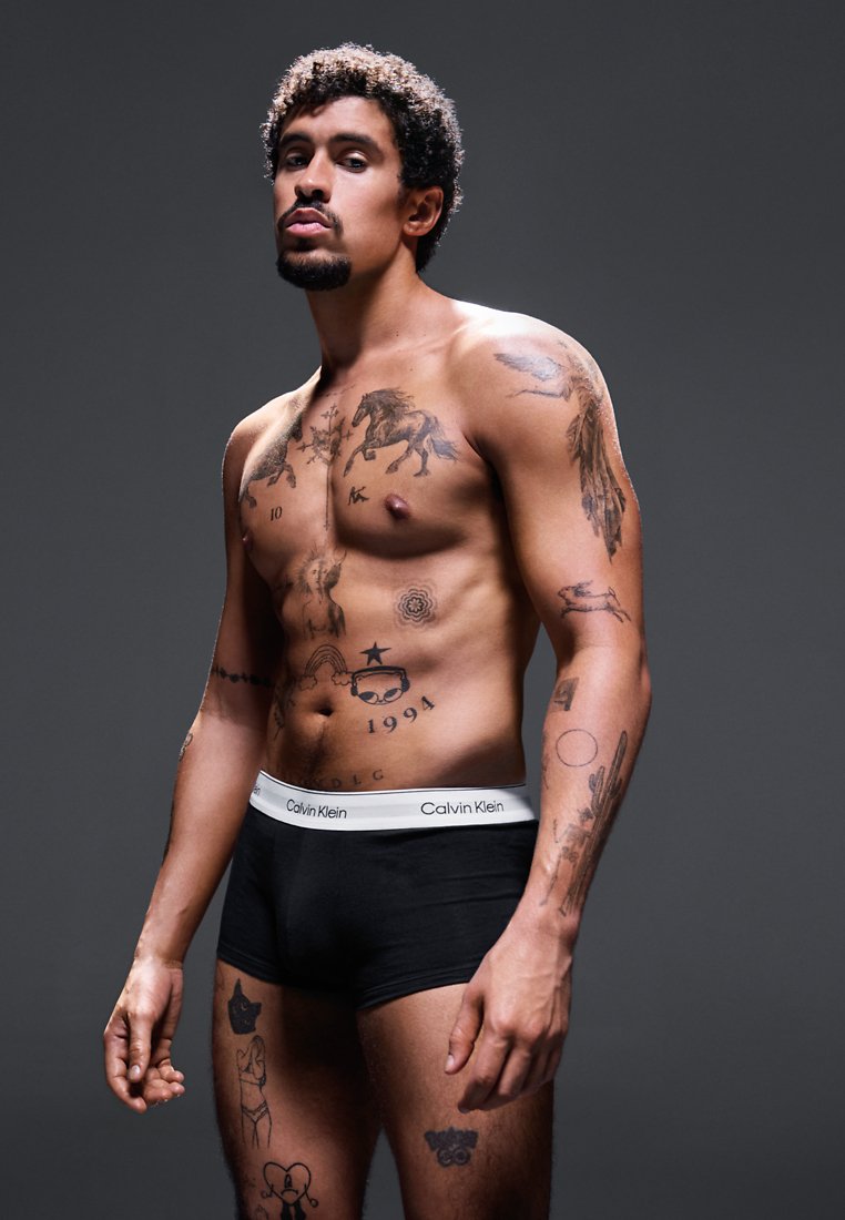 Schwarze Baumwollboxer mit einem weißen Calvin Klein Bund. Das Model hat zahlreiche Tattoos auf seinem Oberkörper und seinen Armen und steht vor einem grauen Hintergrund.